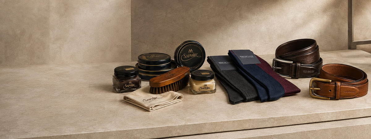 A curated collection of men's grooming accessories arranged on a stone ledge against a textured beige wall. The set includes multiple brown and black leather belts, a brown hairbrush, a folded beige cloth, two small jars with lids, and several folded neckties in navy, burgundy, and black.