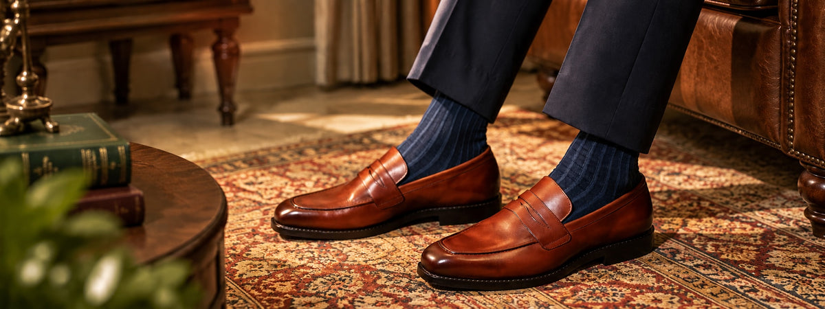A person wearing dark navy trousers, navy socks, and brown leather penny loafers sits on a patterned rug in a warm, traditional room setting.