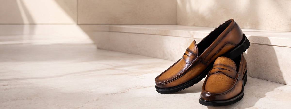 A pair of brown leather penny loafers with dark soles, styled on a light beige stone floor next to a matching stone step.