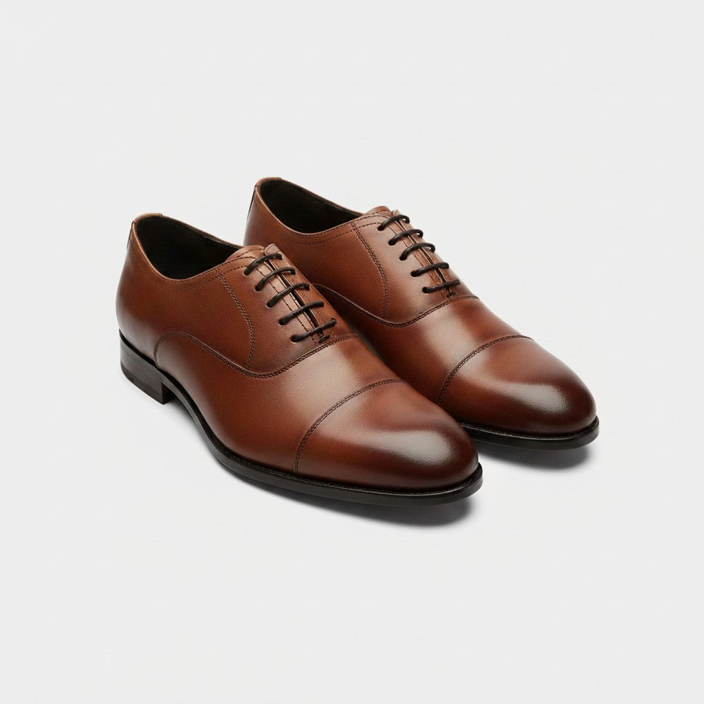 A pair of men's brown leather oxford shoes with cap toes and black laces, displayed on a clean light gray background.